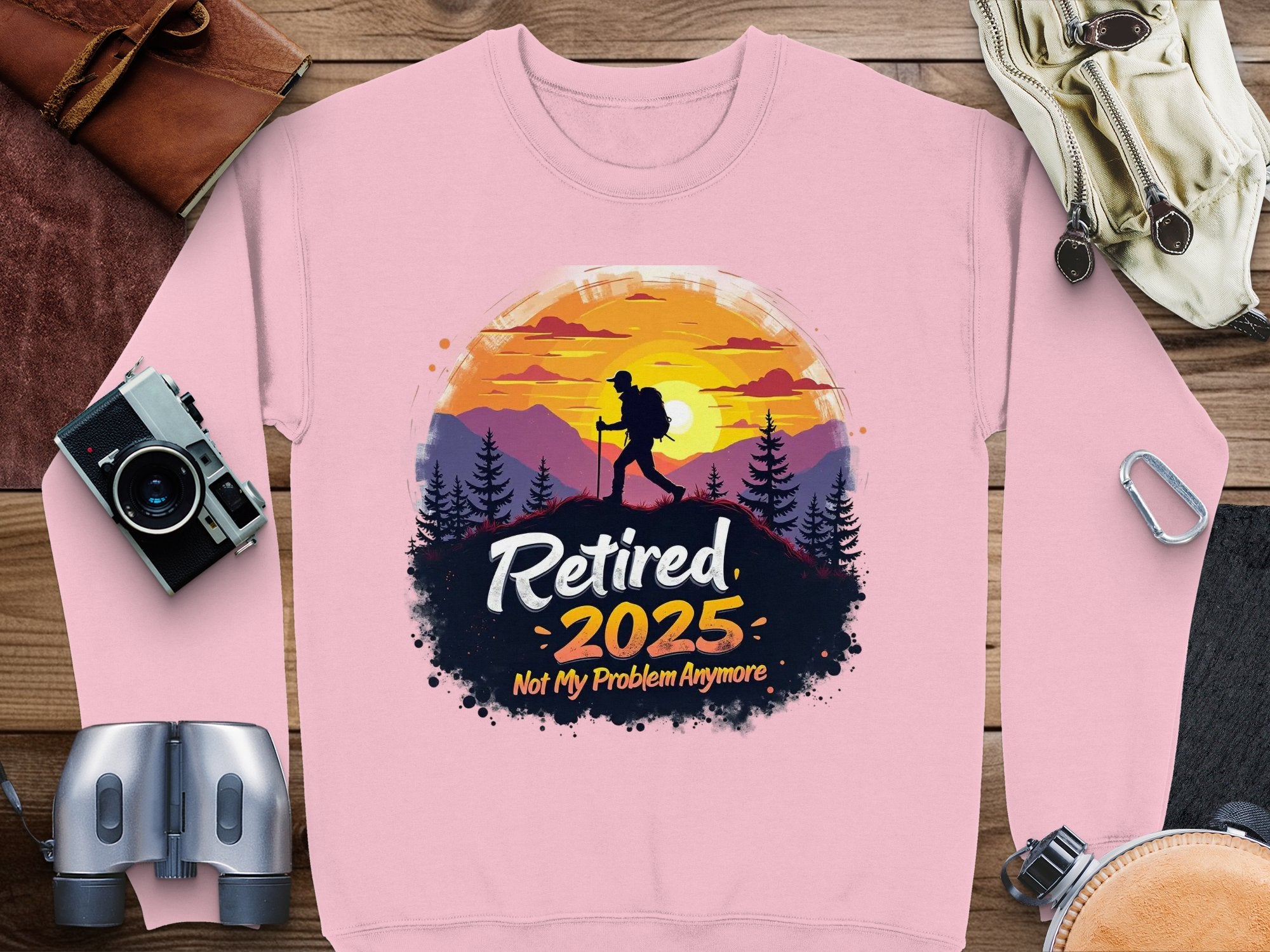 Garment Graphics retirement sweatshirt features a hiker against a sunset encircled by trees, with Retired 2025 Not My Problem Anymore text. Nearby items include a camera, binoculars, map, belt, and bag. It comes in unisex sizing.