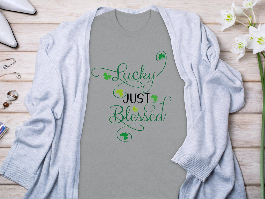 This Royal Mail - 24 Tracked classic fit gray t-shirt displays Lucky Just Blessed in green cursive with small shamrocks. Styled on a wooden surface, its paired with a gray cardigan, flowers, jewelry, and white shoes—an ideal addition to your graphic t-shirt collection.