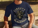 A person sits on a wooden bench wearing a Garment Graphics navy blue T-shirt featuring a skull with goggles. Ideal for motorcycle enthusiasts, it says Liberty Motorcycles Blackpool Specials 2023 and includes wings and wrench graphics, reflecting vintage style with flair.