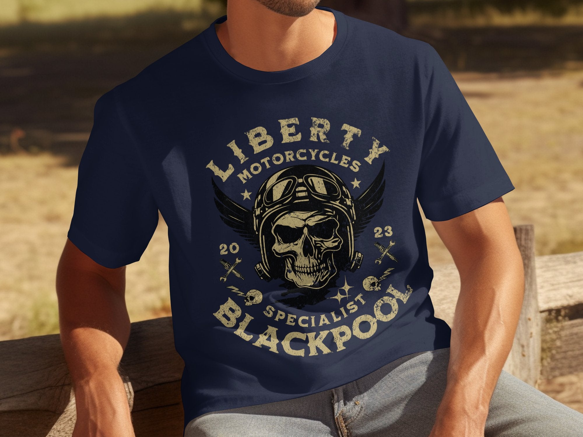 A person sits on a wooden bench wearing a Garment Graphics navy blue T-shirt featuring a skull with goggles. Ideal for motorcycle enthusiasts, it says Liberty Motorcycles Blackpool Specials 2023 and includes wings and wrench graphics, reflecting vintage style with flair.