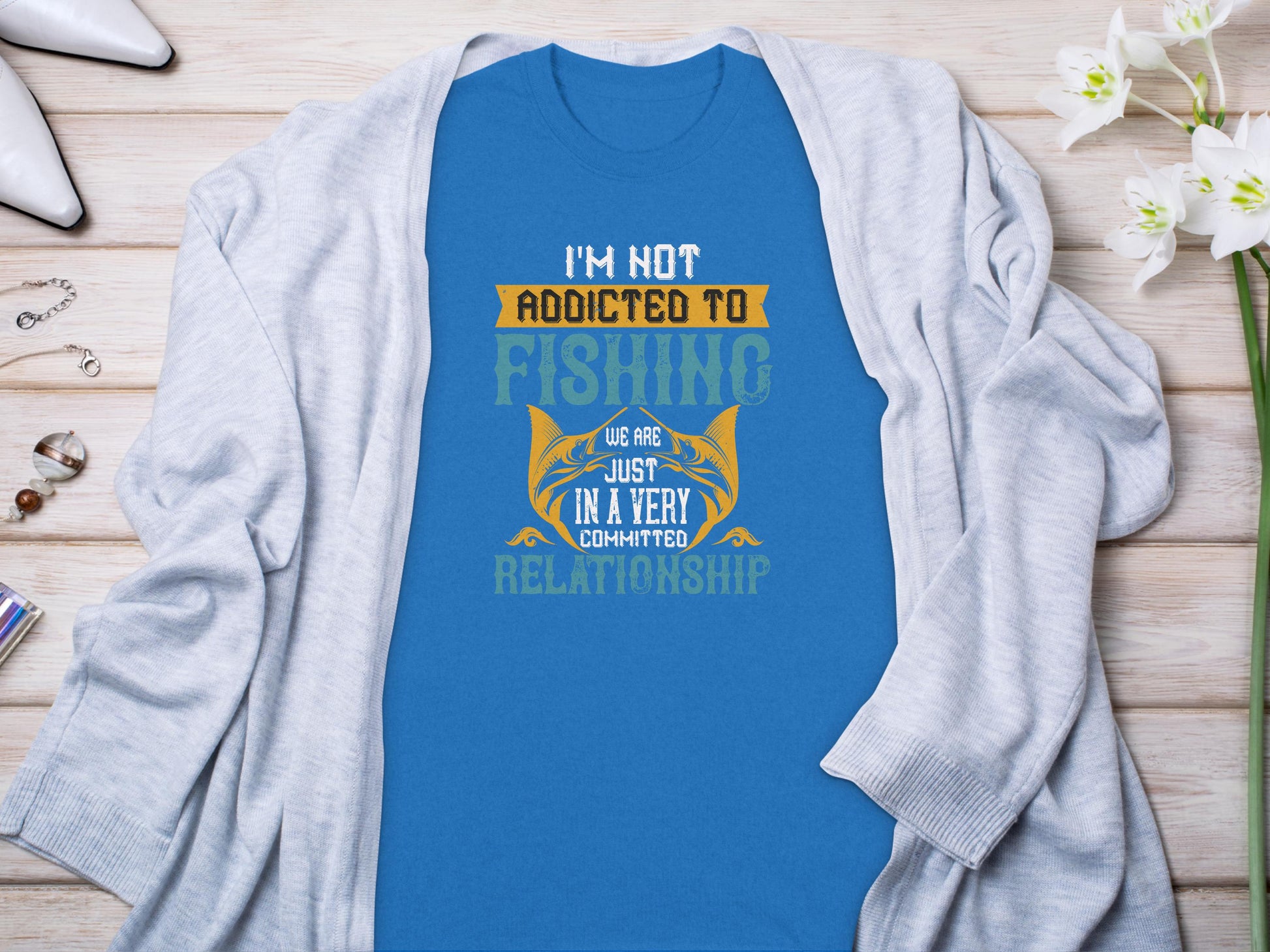 The Blue Fishing T-shirt by Garment Graphics, featuring the text Im not addicted to fishing, we are just in a very committed relationship, is displayed on a light gray cardigan atop a wooden surface, accented with flowers, jewelry, and a watch.