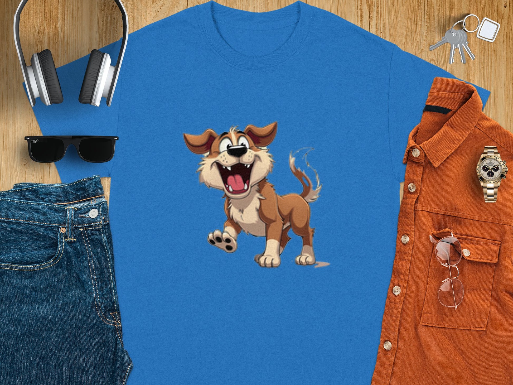 A folded blue cartoon dog t-shirt by Garment Graphics for dog lovers is surrounded by headphones, black sunglasses, denim jeans, a brown shirt with sunglasses, a watch, and keys on a wooden surface.