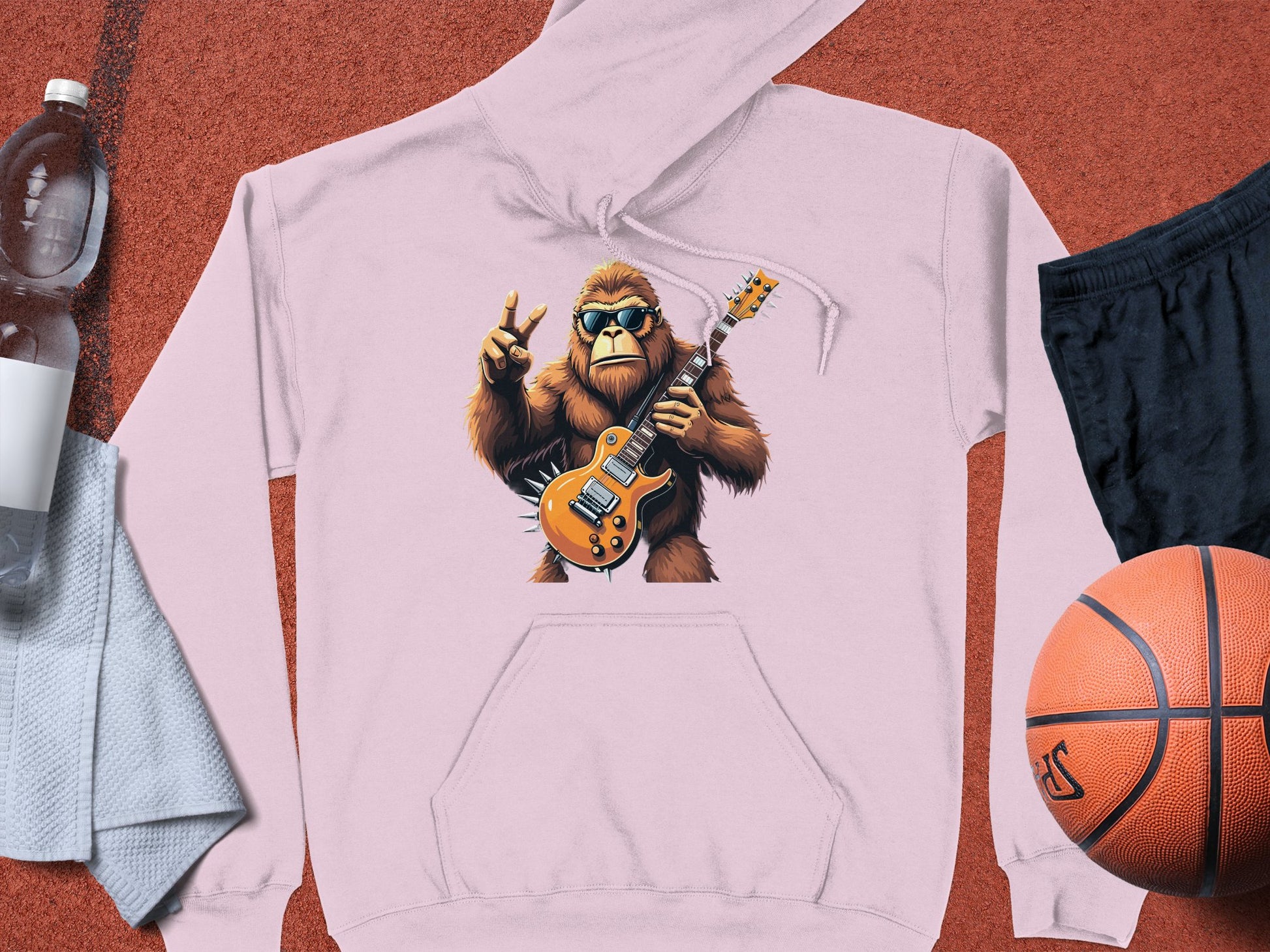 The Garment Graphics Guitar Playing Gorilla Hoodie is perfect for animal and music lovers, showcasing a light pink design with a gorilla holding an electric guitar and making a peace sign. Pair it with essentials like a water bottle, towel, black shorts, and an orange basketball.