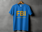 A Garment Graphics blue T-shirt with a funny design hangs on a wooden hanger against a gray wall. In bold yellow, it reads FBI with smaller text below saying Female Boob Inspector.
