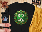 A Garment Graphics black shirt with a green shamrock design, celebrating the Irish spirit, is displayed on a wooden surface. Nearby are a yellow jacket, makeup palette, sunglasses, makeup brushes, pink shoes, nail polish, and a small blue pouch.