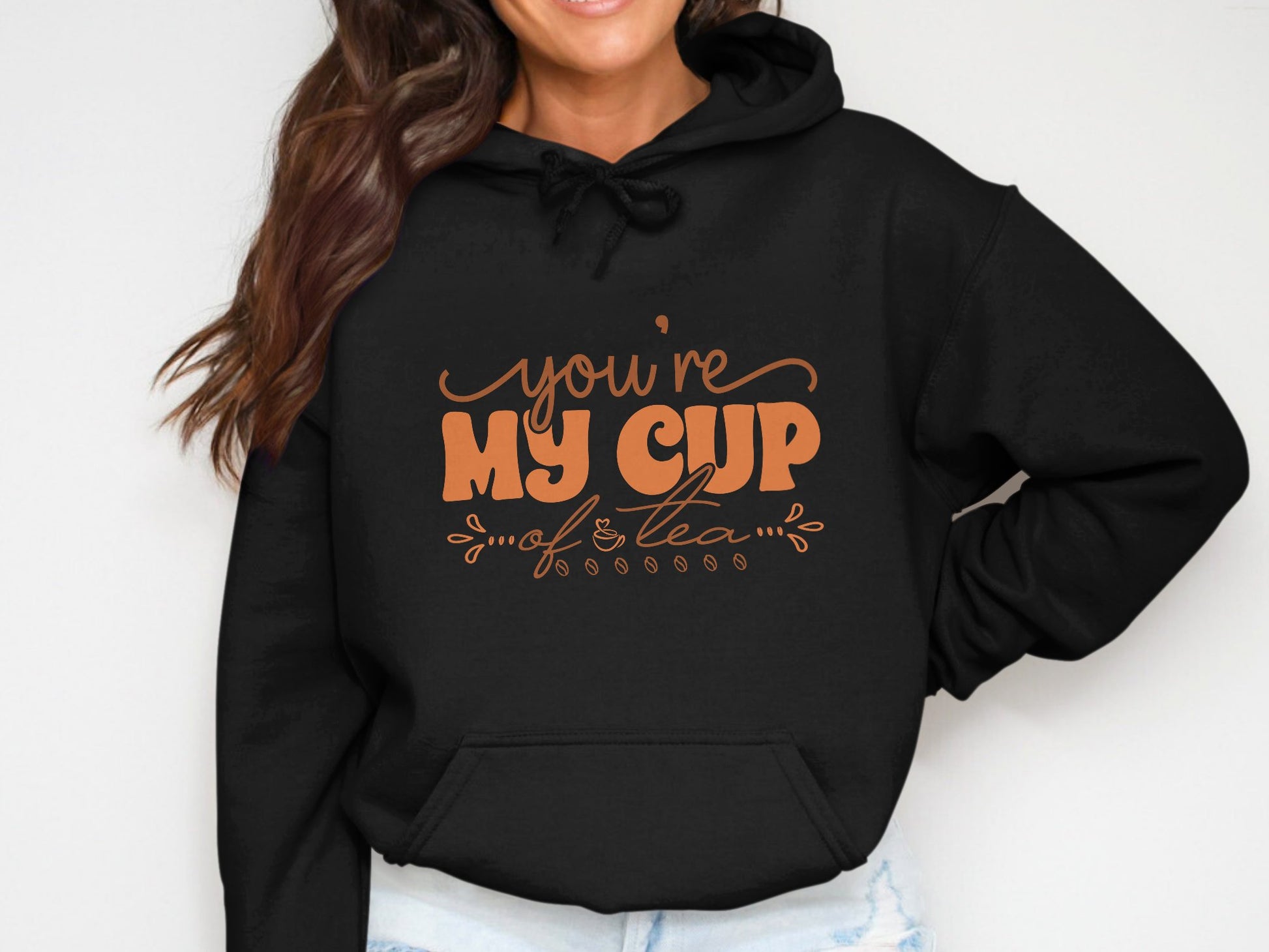 A person confidently wearing a Garment Graphics black hoodie with an orange youre my cup of tea design stands with hands on hips. Their long, wavy hair complements the cozy moment, accented by the hoodies snug drawstring neckline.