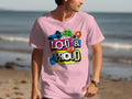 A person is wearing a light pink Garment Graphics T-shirt featuring a colorful Hey Ho Let’s Go Loud & Proud design in varied fonts. They stand on the beach, looking aside as the ocean and sandy shore provide an ideal backdrop, highlighting their passion.