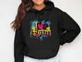 The individual sports a Garment Graphics black hoodie with a vibrant graffiti design, featuring I love graffiti text with colorful splashes and brick motifs, embodying urban street art. With long hair and a smile, their casual style is completed by hands tucked in pockets.