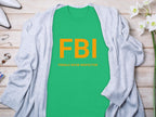 A Garment Graphics green shirt with FBI in bold yellow, reading Female Boob Inspector, is laid flat on wood. Its surrounded by a gray cardigan, white flowers, a ring, bracelet, and stylish white shoes, epitomizing humorous t-shirts.