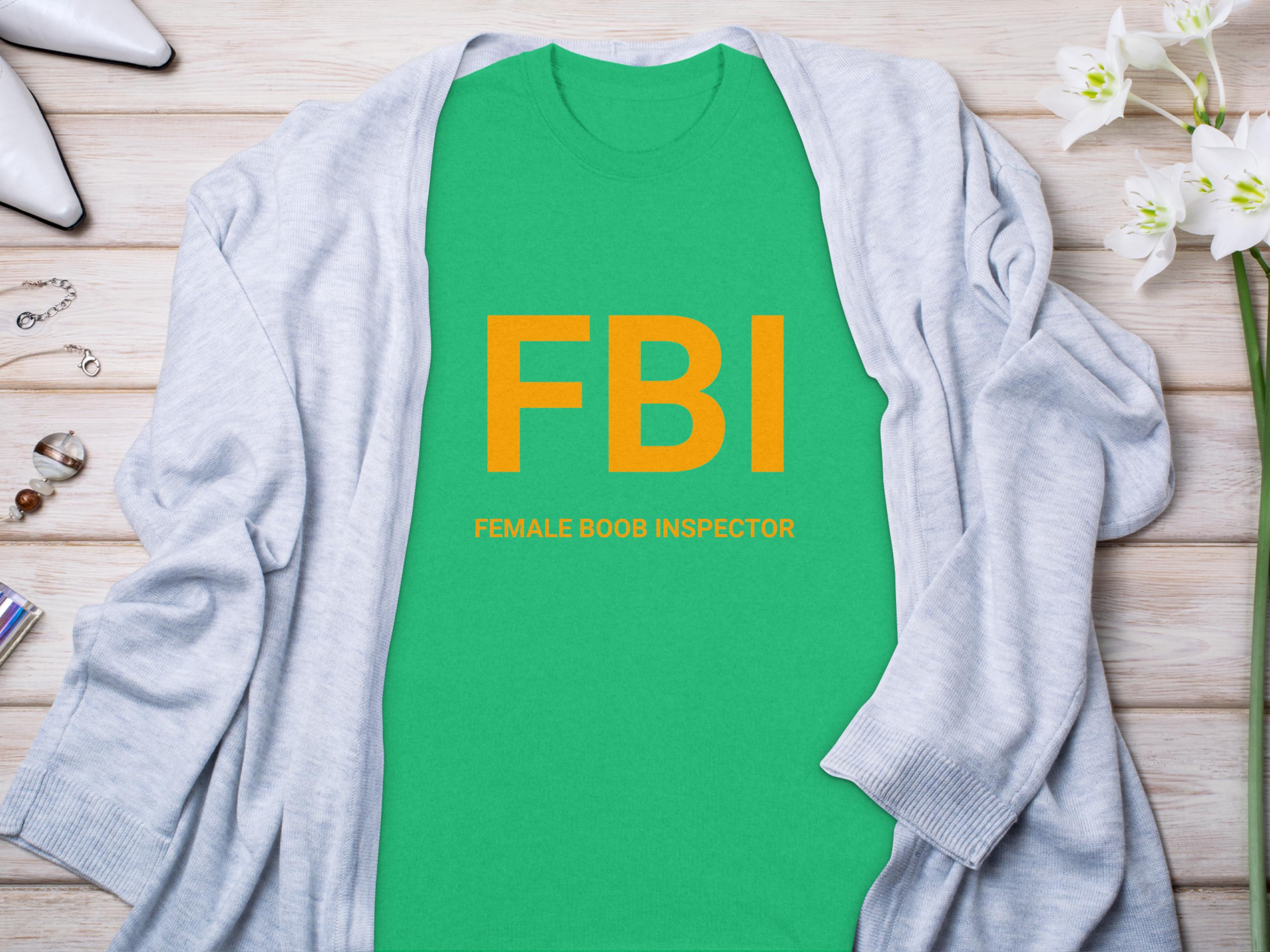 A Garment Graphics green shirt with FBI in bold yellow, reading Female Boob Inspector, is laid flat on wood. Its surrounded by a gray cardigan, white flowers, a ring, bracelet, and stylish white shoes, epitomizing humorous t-shirts.