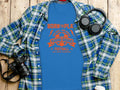 A Garment Graphics blue football T-shirt with Born to Play and Young Squad Football printed on it, set against a wooden surface next to a plaid shirt, headphones, and a camera, epitomizing the pursuit of that one goal and dream.