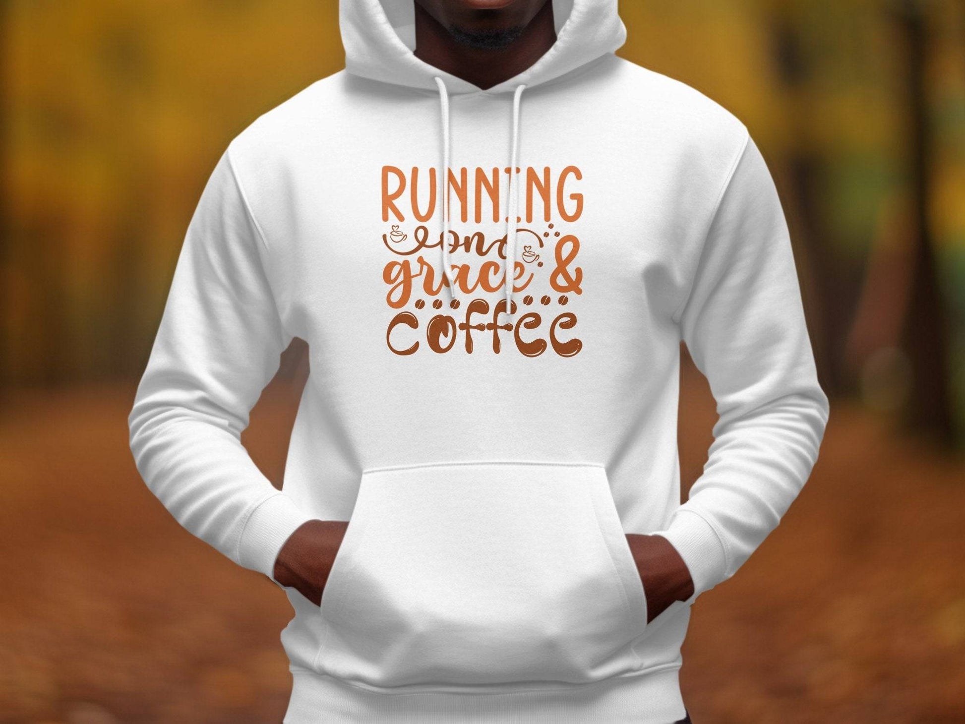 A person in a Garment Graphics medium-heavy classic fit hoodie stands outside with Running on Grace & Coffee in warm brown and orange letters, beautifully matching the blurred autumn foliage.