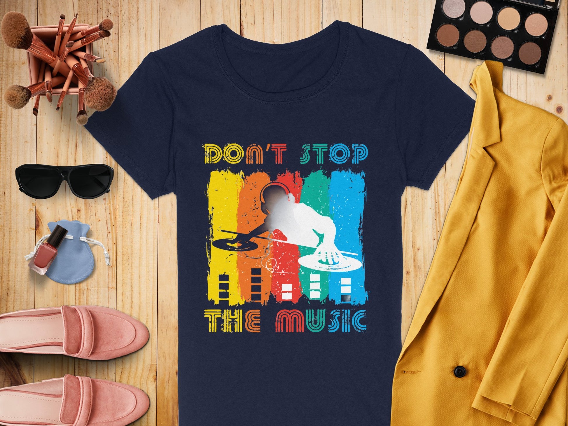 A Garment Graphics dark blue DJ graphic tee features Dont Stop the Music and a turntable design, perfect for music enthusiasts. Its paired with vibrant accessories like makeup brushes, sunglasses, nail polish, shoes, a makeup palette, and a yellow blazer on a wooden surface.
