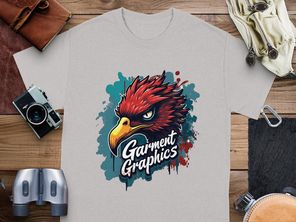 A gray classic fit tee features a vibrant eagle design with Garment Graphics underneath, placed on a wooden table surrounded by a leather pouch, gloves, vintage camera, binoculars, and bracelet.