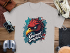 A gray classic fit tee features a vibrant eagle design with Garment Graphics underneath, placed on a wooden table surrounded by a leather pouch, gloves, vintage camera, binoculars, and bracelet.