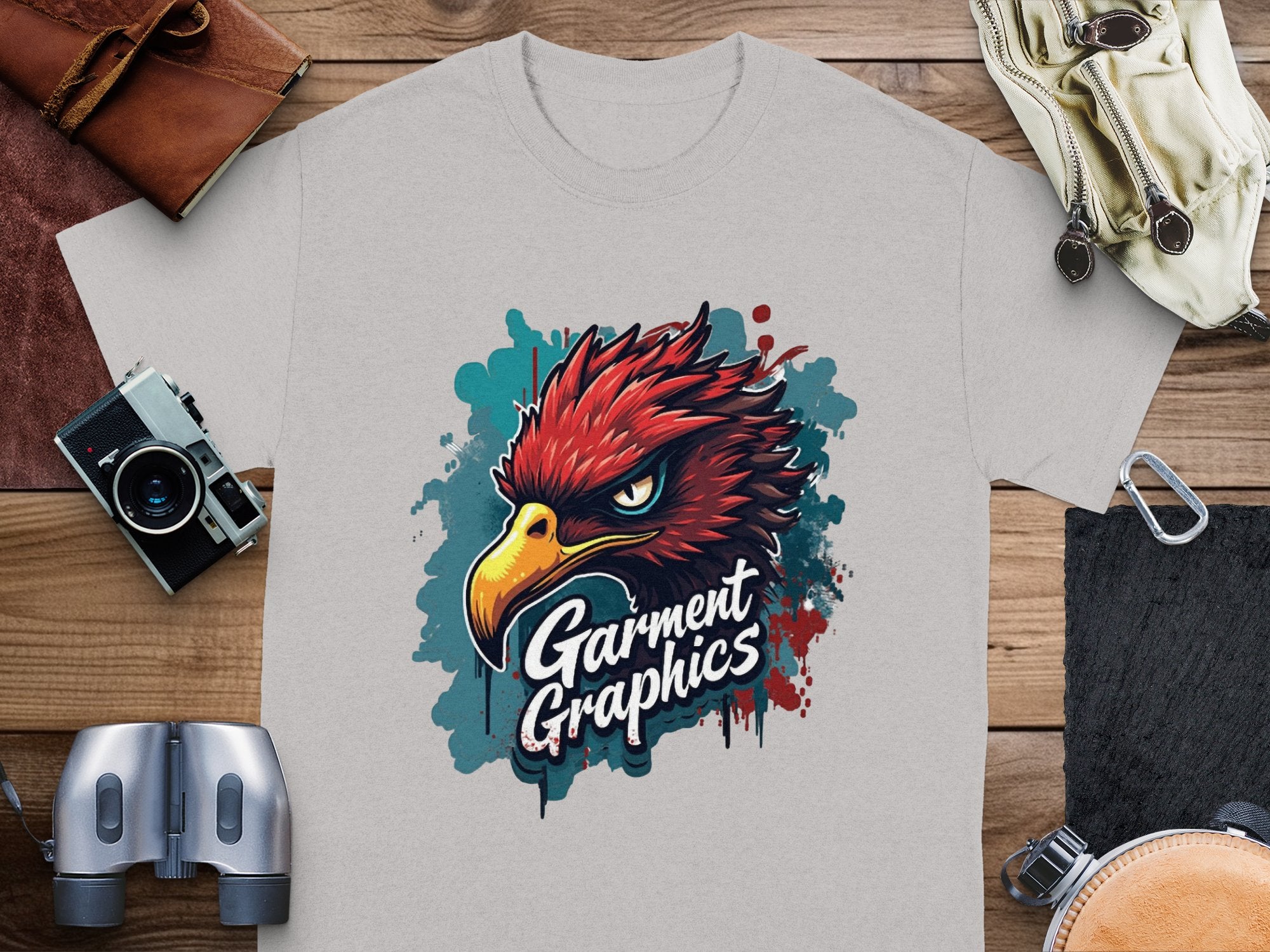 A gray classic fit tee features a vibrant eagle design with Garment Graphics underneath, placed on a wooden table surrounded by a leather pouch, gloves, vintage camera, binoculars, and bracelet.