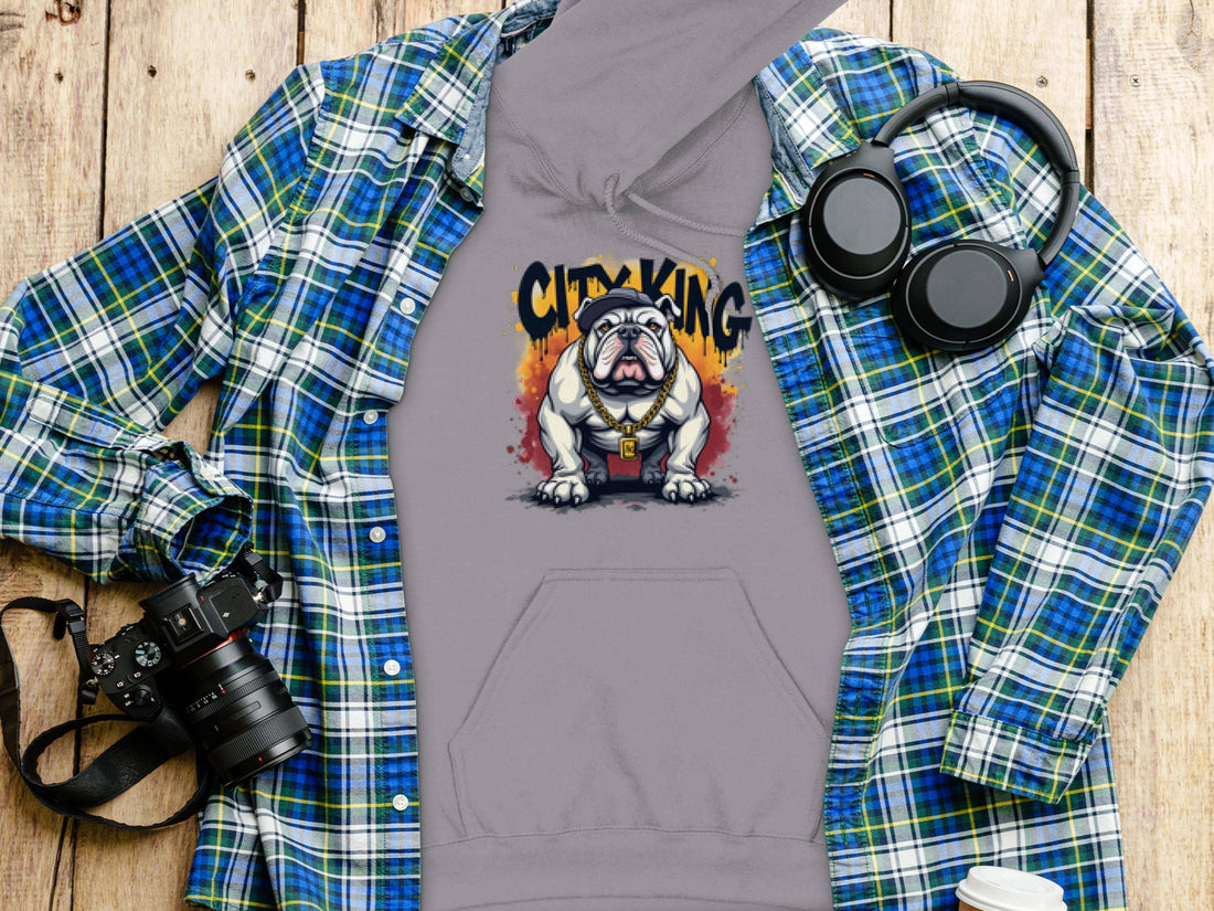 A Garment Graphics gray hoodie with a Graphic Bulldog Design and CITY KING text rests on a wooden surface. Ideal for casual wear, its accompanied by a blue plaid shirt, camera, headphones, and a white lidded cup.