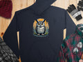 The Garment Graphics Fierce Owl Illustration Hoodie, crafted from medium-heavy fabric, boasts a unique style. Displayed on a wooden surface with winter essentials like hat, gloves, plaid shirt, and scarf, its owl design features a standout circular background.