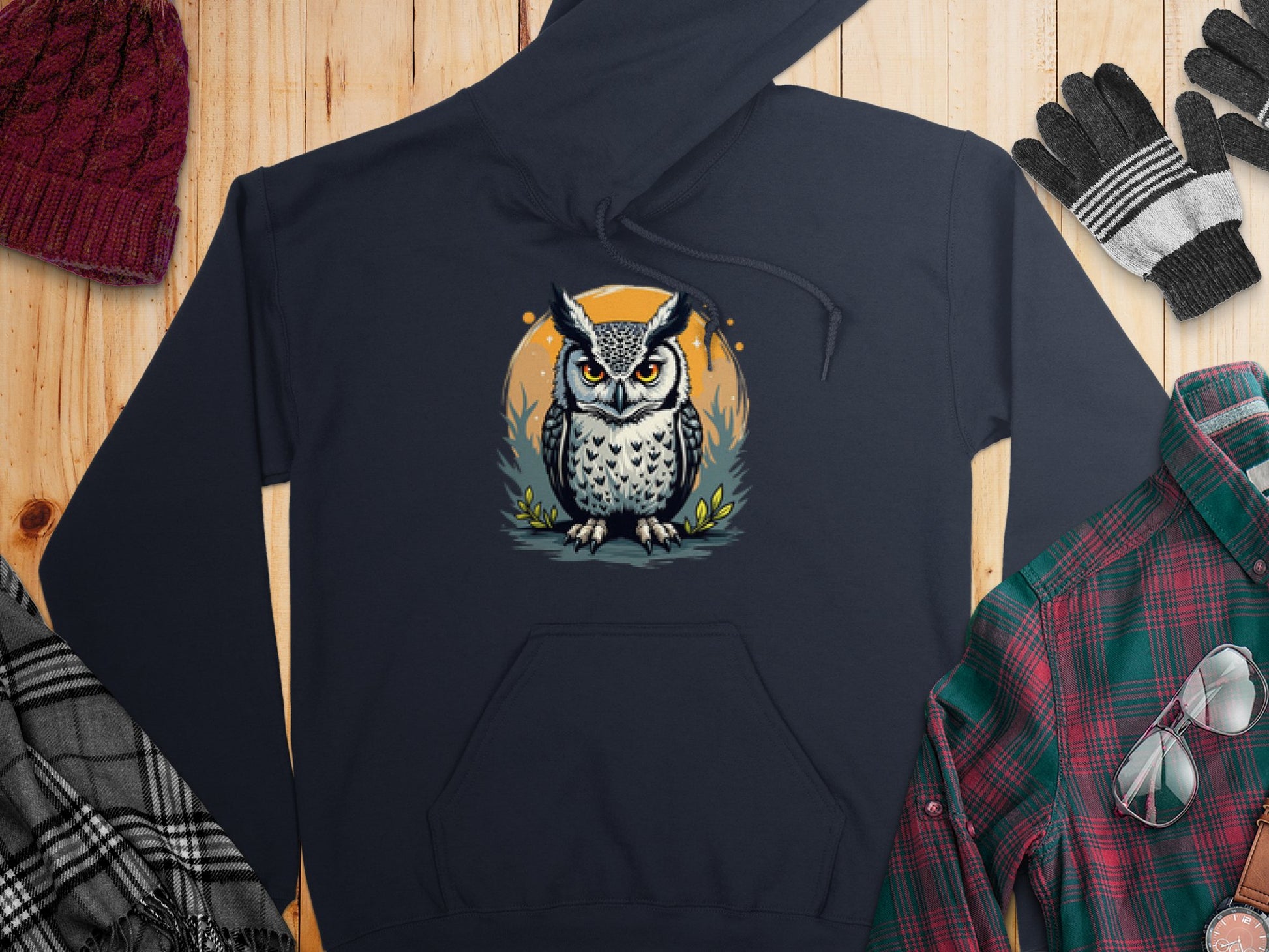 The Garment Graphics Fierce Owl Illustration Hoodie, crafted from medium-heavy fabric, boasts a unique style. Displayed on a wooden surface with winter essentials like hat, gloves, plaid shirt, and scarf, its owl design features a standout circular background.