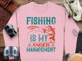 A stylish pink sweatshirt from Garment Graphics features Fishing Is My Anger Management in green and orange letters with a fish and lure design, ideal for fishing fans. Its pictured surrounded by a camera, binoculars, and leather strap on a wooden surface.