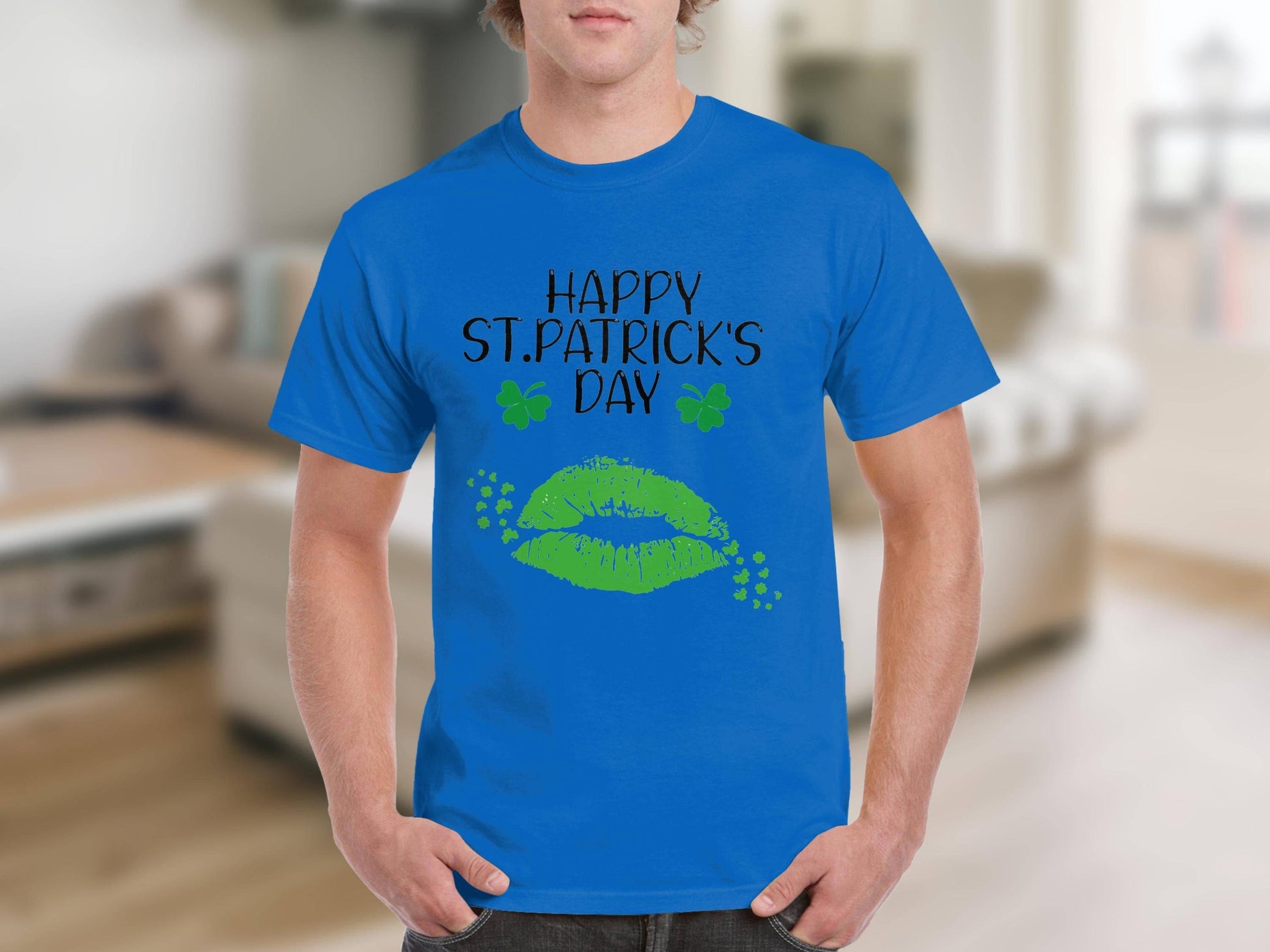 A person sports a Garment Graphics T-shirt featuring Happy St. Patricks Day in black, accented by green lips and clovers, against a blurred indoor backdrop with furniture for a festive vibe.