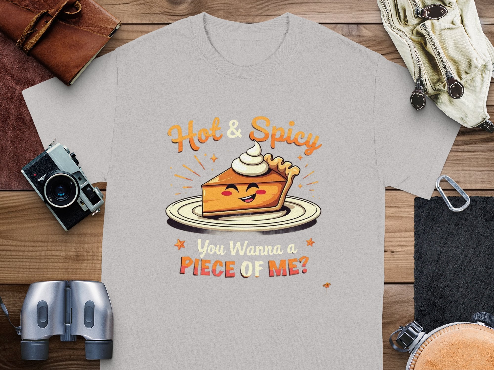 The Hot and Spicy Pie T-shirt by Garment Graphics displays a cartoon pie slice with sunglasses and whipped cream, surrounded by orange stars, alongside the playful text You Wanna a Piece of Me? Set against items like a camera, it is available in family sizes.
