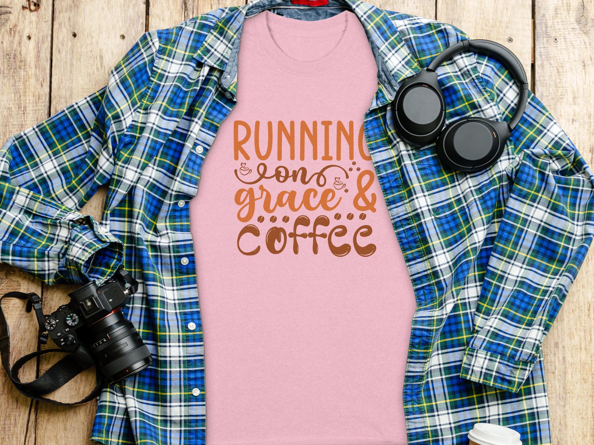 Garment Graphics pink Running on Grace & Coffee t-shirt is styled over a blue and green plaid shirt, set on a wooden surface. Accompanied by a black camera, headphones, and a white cup, this scene perfectly blends inspiration with style.