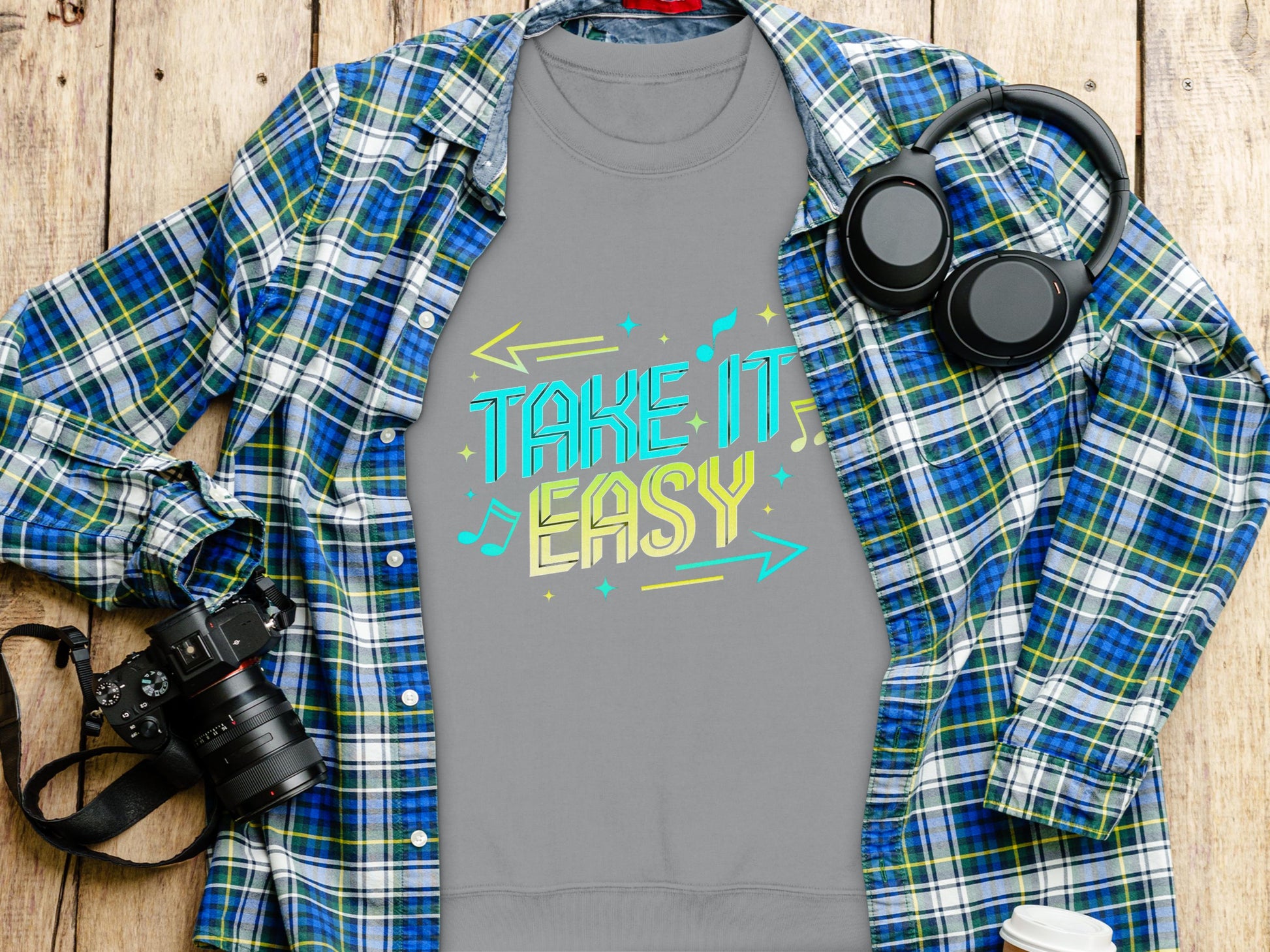 A Garment Graphics gray T-shirt featuring the colorful Take It Easy design lays on wooden surface. In unisex sizes, it pairs perfectly with a blue and green plaid shirt, while a camera, headphones, notebook, and coffee cup complete the cozy scene.