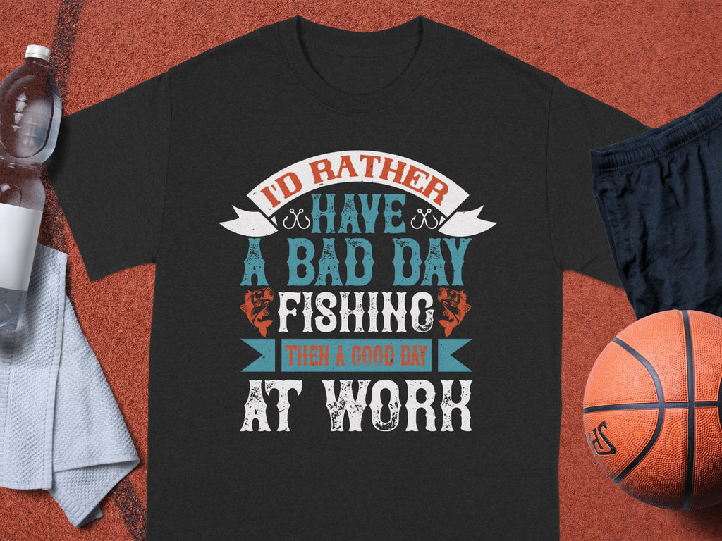 A black T-shirt from Garment Graphics features the text Id rather have a bad day fishing than a good day at work in stylized fonts. Ideal for anglers, its displayed on a brown surface alongside a water bottle, towel, black shorts, and basketball.