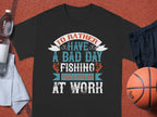 A black T-shirt from Garment Graphics features the text Id rather have a bad day fishing than a good day at work in stylized fonts. Ideal for anglers, its displayed on a brown surface alongside a water bottle, towel, black shorts, and basketball.