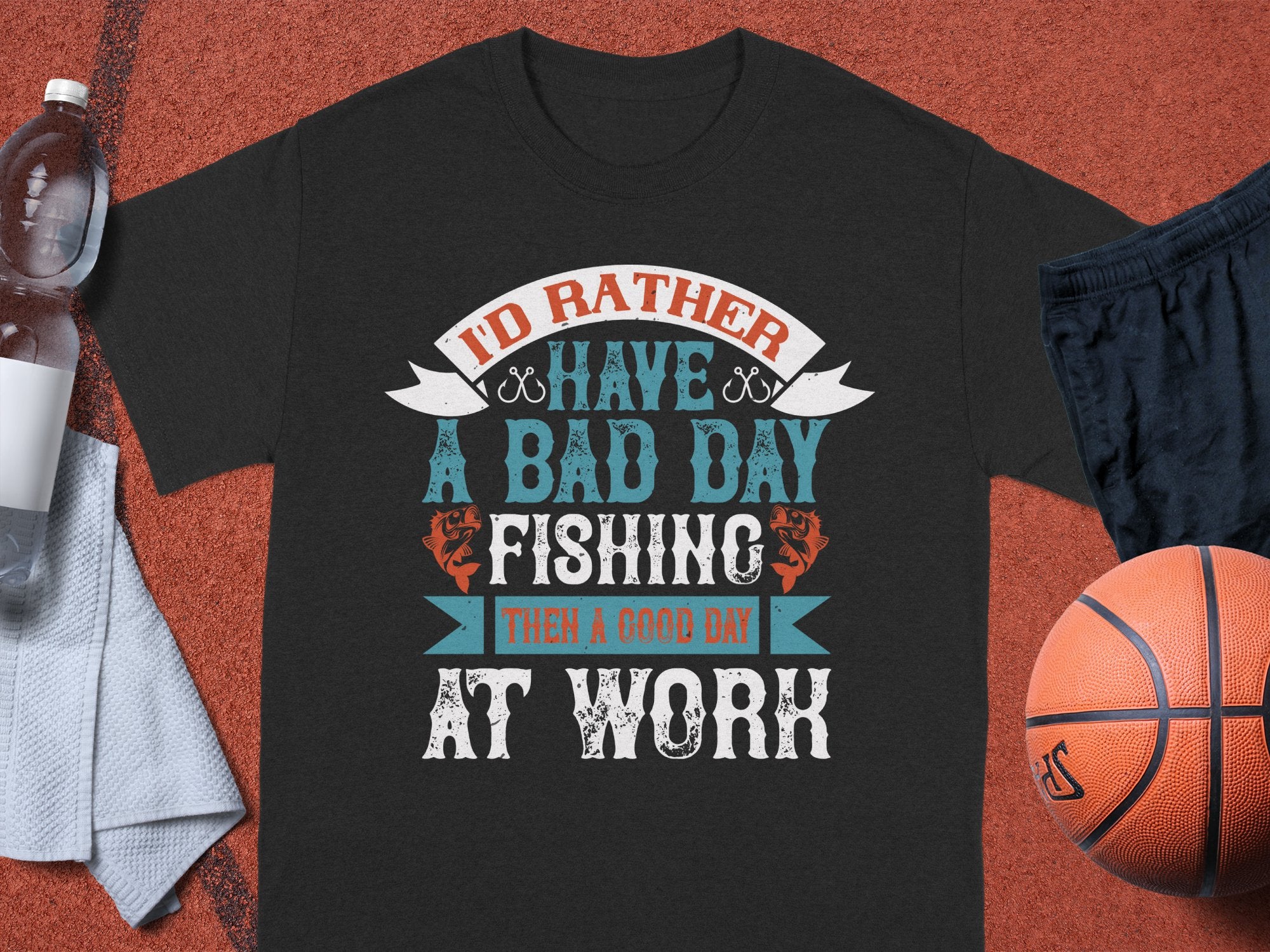 A black T-shirt from Garment Graphics features the text Id rather have a bad day fishing than a good day at work in stylized fonts. Ideal for anglers, its displayed on a brown surface alongside a water bottle, towel, black shorts, and basketball.