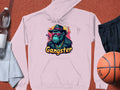 Garment Graphics light pink hoodie captures hipster urban style with a graphic of a hat-wearing, sunglasses-clad gangster ape and the word Gangster below. Set with a basketball, black shorts, towel, and two water bottles, its ideal for any casual outing.