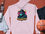 Garment Graphics light pink hoodie captures hipster urban style with a graphic of a hat-wearing, sunglasses-clad gangster ape and the word Gangster below. Set with a basketball, black shorts, towel, and two water bottles, its ideal for any casual outing.