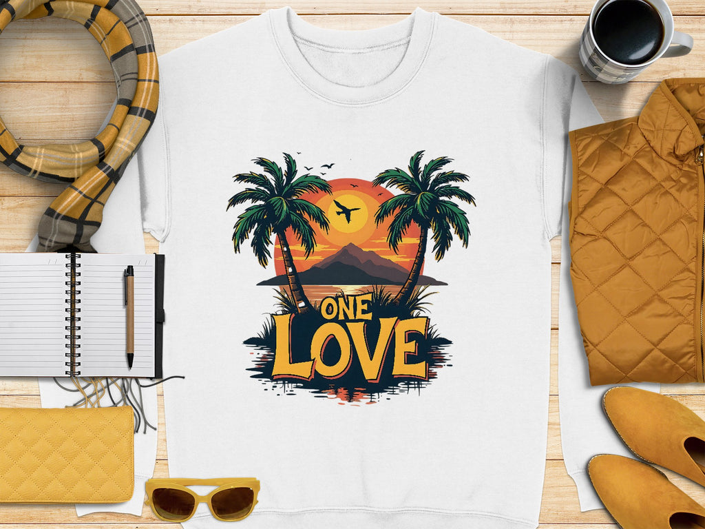The One Love Sunset white T-shirt by Garment Graphics showcases palm trees framing a sunset with birds and mountains, surrounded by a notepad, pen, yellow purse, sunglasses, scarf, and coffee cup.