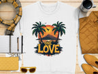 The One Love Sunset white T-shirt by Garment Graphics showcases palm trees framing a sunset with birds and mountains, surrounded by a notepad, pen, yellow purse, sunglasses, scarf, and coffee cup.