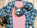 A stylish pink T-shirt by Garment Graphics features a striking wolf graphic laid out on a wooden surface, partially covered by a blue and green checkered shirt. Nearby are a black camera, headphones, and a white bottle, exuding fearless personality.