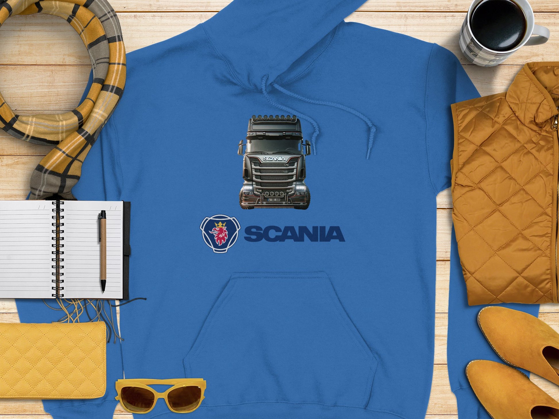 The Blue Scania Truck Hoodie by Garment Graphics, with a graphic print and logo, is stylishly arranged on a wooden surface with a notebook and pen, yellow sunglasses, quilted clutch, checkered scarf, yellow jacket, and coffee cup—ideal for truck enthusiasts.