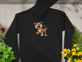 A Garment Graphics black cartoon puppy hoodie, showcasing a playful laughing dog design, lies flat on concrete. This fun piece for dog lovers is adorned with green plants around it, a red flower to the left, and yellow flowers to the right.