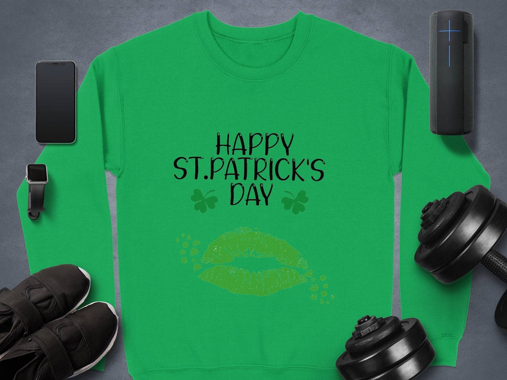 A Garment Graphics green sweatshirt with Happy St. Patricks Day and clovers in black print, featuring a cheeky clover kiss. It’s surrounded by a smartphone, fitness tracker, shoes, dumbbells, and a portable speaker on gray—ideal for capturing the festive spirit.