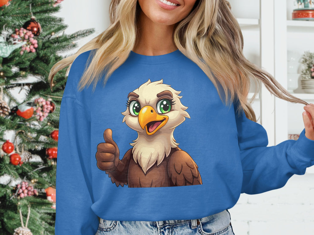 A person wears a cheerful blue cartoon eagle sweatshirt from Garment Graphics, featuring an eagle giving a thumbs up. They stand beside a festively decorated Christmas tree and an ornament-adorned white shelf. The unisex sizing suits different styles, though their face is not visible.