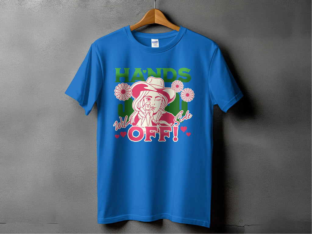 Garment Graphics presents a blue T-shirt on a hanger with a bold design of a woman in a hat, finger to lips, surrounded by flowers. It features HANDS OFF! text and Wild Side in smaller print with hearts and stars. Available in family sizes for all.