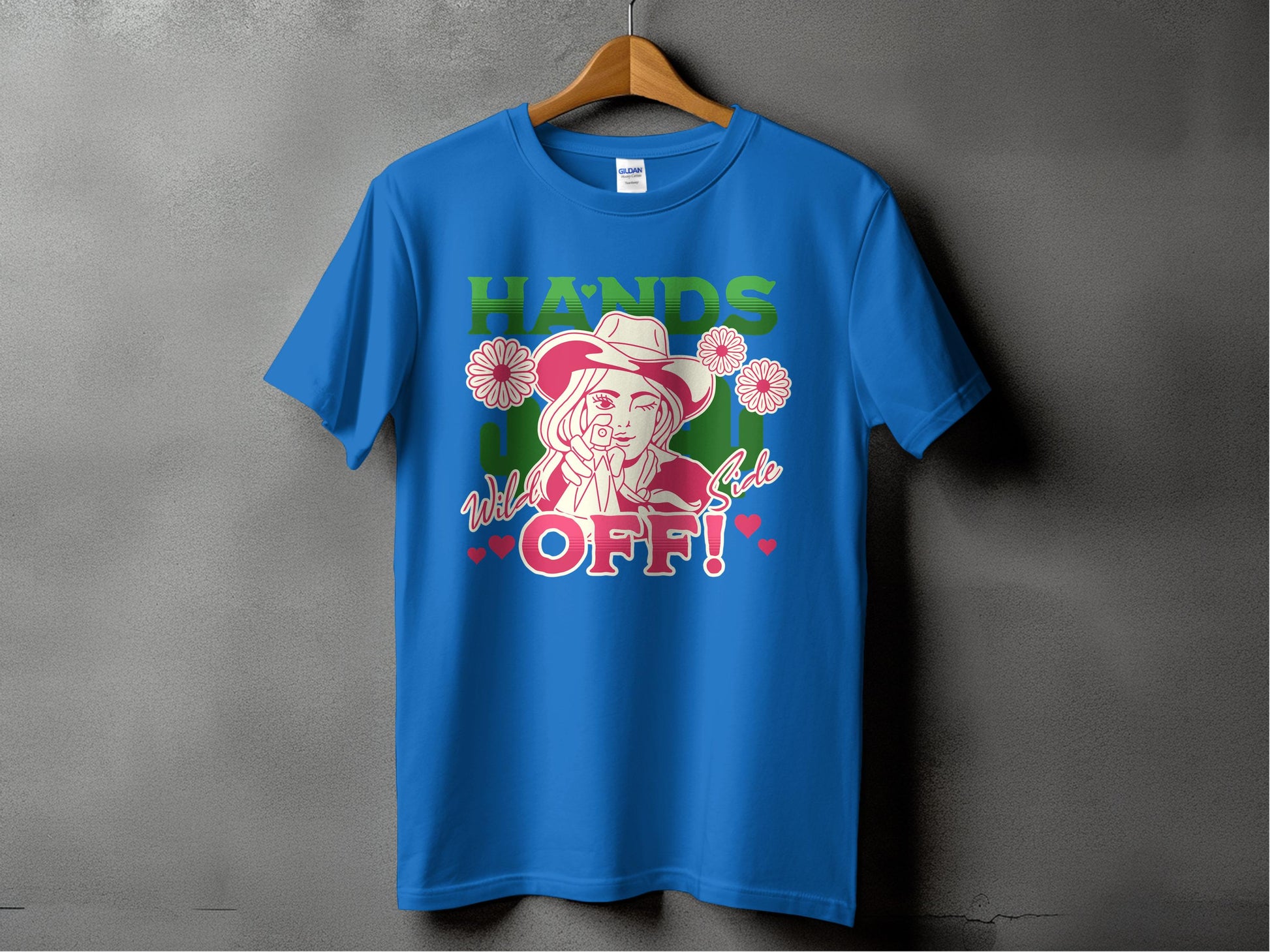 Garment Graphics presents a blue T-shirt on a hanger with a bold design of a woman in a hat, finger to lips, surrounded by flowers. It features HANDS OFF! text and Wild Side in smaller print with hearts and stars. Available in family sizes for all.