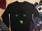 The black festive sweatshirt by Garment Graphics features Happy St. Patricks Day in black letters, adorned with green shamrocks and a heart. It stands out beautifully on a wooden surface, paired with a red hat, gloves, and plaid shirts nearby.
