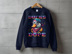 A navy blue Garment Graphics sweatshirt hangs on a wooden hanger against a concrete wall, featuring a cartoon duck with glasses and a hat, and DUCKS DOPE in colorful letters. Made from medium-heavy fabric, it offers unisex sizing for the perfect fit.