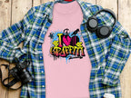 A flat lay showcases a pink T-shirt by Garment Graphics with an I Love Graffiti design, paired with an urban blue plaid shirt. Nearby items like a camera, headphones, and a white bottle rest on wood, capturing the street art vibe.