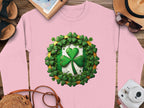 A Garment Graphics pink sweatshirt with a green clover motif and smaller clovers embodies Irish spirit. Nearby, sandals, a brown hat, sunglasses, a white camera, and a notebook with pen sit on a wooden surface.
