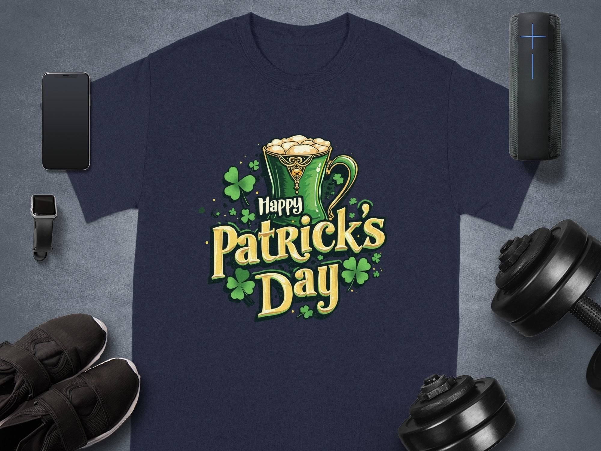 Garment Graphics navy statement St. Patricks Day t-shirt features a green mug, shamrocks, and Happy Patricks Day, artistically framed by a smartphone, watch, shoes, speaker, and dumbbells on a sleek gray background.