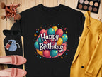 Garment Graphics presents a stylish black shirt with colorful balloons and Happy Birthday text, perfect for celebrations. Its paired with a yellow blazer, pink shoes, makeup products, sunglasses, and a small blue pouch on the wooden surface. Available in unisex sizes for all!.