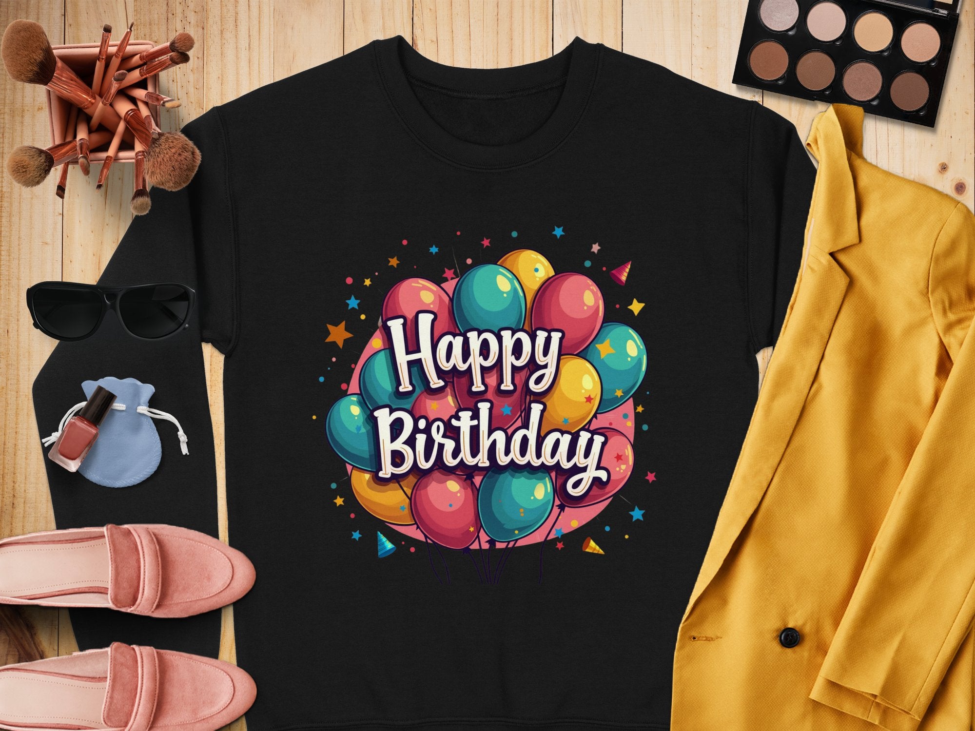 Garment Graphics presents a stylish black shirt with colorful balloons and Happy Birthday text, perfect for celebrations. Its paired with a yellow blazer, pink shoes, makeup products, sunglasses, and a small blue pouch on the wooden surface. Available in unisex sizes for all!.