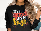 A person with long, wavy hair wears a Garment Graphics sweatshirt made from medium-heavy fabric. The black design features colorful text, Its a good day to laugh, with red, yellow, and white splatters, available in unisex sizes for everyone to enjoy.